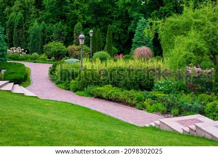 stone steps descent to a path from paving slabs in park with iron retro lantern on slope with scenic plants, evergreen bushes and pine and foliage trees, backyard landscape place to be at peace. Royalty-Free Stock Photo #2098302025