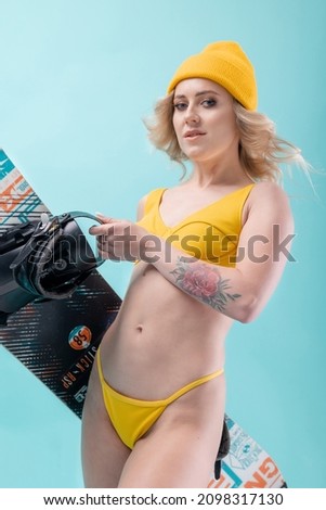 A sporty blonde with curly hair, in a yellow bikini swimsuit, in a yellow hat, posing on a blue background with a snowboard. Studio, flash.