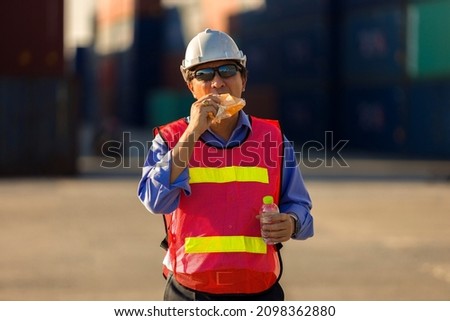 An Asian engineer or container yard worker in protective clothing stands for a snack during the break. Concept of safety and happiness at work.