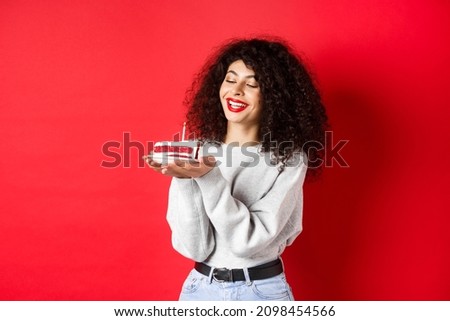 Happy birthday girl celebrating and making wish, holding bday cake and smiling, standing on red background