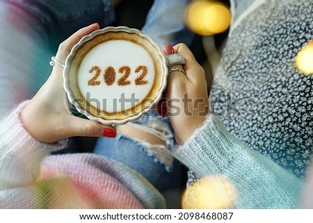 Holidays food art number 2022 on frothy surface of cappuccino served in white cup holding by woman in pastel rainbow knitted sweater with jeans relaxed sitting on the couch. Happy New Year 2022 theme.