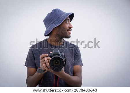 Professional photographer holds a camera in hand. Stylish and attractive man wears a bucket hat and smiles while holding a DSLR, he is isolated on white background