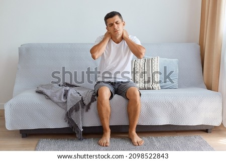 Portrait of ill unhealthy man wearing white T-shirt sitting on cough in living room, touching or massaging his painful neck, uncomfortable sleeping conditions, vertebral column injury. Royalty-Free Stock Photo #2098522843