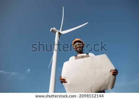 African american man in uniform, helmet and glasses working with blueprints among windmill farm. Professional technician checking and maintaining production of green energy.