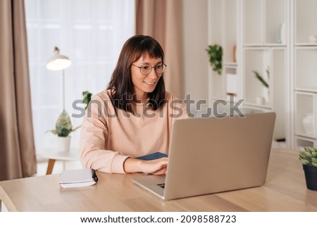 Pretty smiling woman in glasses working on laptop in home office, sitting at desk. Female freelancer working remotely. Student using computer for distance learning or retraining, typing on keyboard Royalty-Free Stock Photo #2098588723