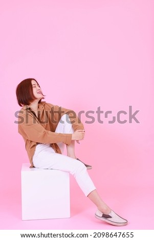 Portrait studio cutout shot of Asian young pretty short hair female model in long brown coat jacket casual shoes sitting crossed legs smiling look at camera on box chair posing on white background.