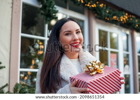 Christmas concept. Beautiful young dark haired woman in beige eco coat opens red gift box with bow. Stylish model is shopping in European city during the winter holidays.