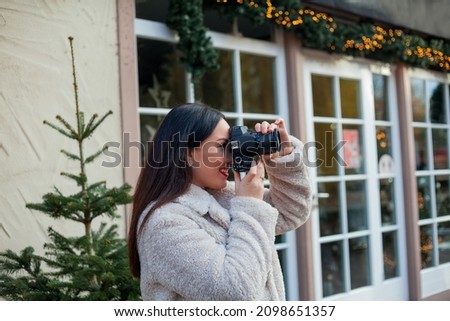 Beautiful young dark haired woman tourist in beige eco-coat makes Christmas photo on European street. Stylish model takes pictures with professional camera. Winter holidays.
