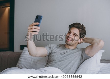 Calm young man in pajamas grey t-shirt lying sit in bed do selfie shot on mobile cell phone post on social network rest relax at home indoors bedroom. Good mood sleeping night morning bedtime concept.