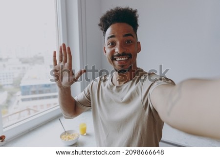 Close up smiling happy man of African American ethnicity in casual clothes wave hand doing selfie shot pov on mobile phone prepare cook food in light kitchen at home alone indoor. Healthy diet concept