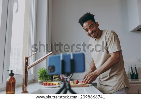Young man of African American ethnicity in casual clothes prepare sweet breakfast cut fruit do selfie shot on mobile cell phone cook food in indoor kitchen at home alone Healthy diet lifestyle concept