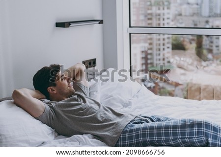 Side viw calm peaceful minded young man 20s wearing pajamas grey t-shirt lying in bed sleep slumber resting look at window relaxing at home indoors bedroom. Good mood night morning bedtime concept. Royalty-Free Stock Photo #2098666756