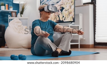 Old woman wearing vr glasses and doing meditation while sitting in lotus position on yoga mat. Senior person using virtual reality goggles to meditate for wellness and recreation. Royalty-Free Stock Photo #2098680904