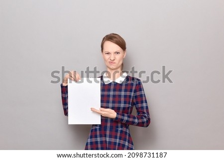 Studio portrait of disgruntled young woman holding white blank paper sheet, pointing with palm at empty place for your text and design, wearing checkered dress, standing over gray background