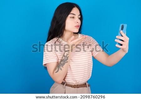 Caucasian girl wearing striped T-shirt over blue background blows air kiss at camera of smartphone and takes selfie, sends mwah via online call.