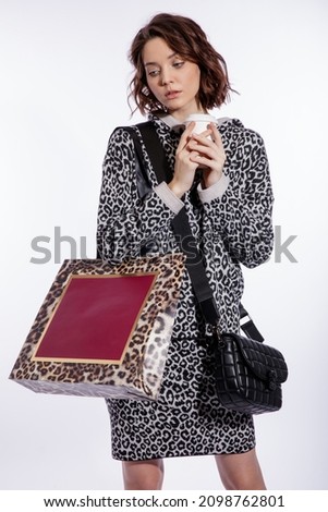 High fashion photo of a beautiful elegant young woman in a pretty gray leopard print blouse, skirt, black handbag posing over white background. Walking style. Hairstyle. Slim figure. Red shopping bag