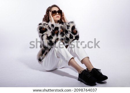 High fashion photo of a beautiful elegant young woman in a pretty leopard print fur coat, stylish sunglasses, trousers, pants posing on white background. Slim figure. Studio shot. Model is sitting