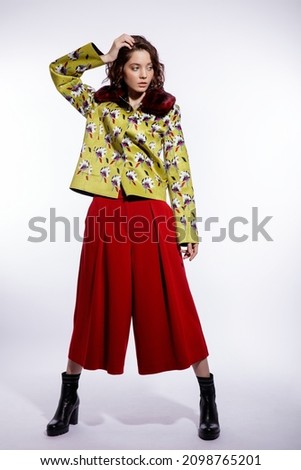 High fashion photo of a beautiful elegant young woman in a pretty yellow jacket with pattern flowers, red trousers, pants posing over white background. Make up, hairstyle. Slim figure. Studio shot