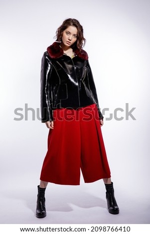 High fashion photo of a beautiful elegant young woman in a pretty black leather jacket, red trousers, pants posing over white background. Make up, hairstyle. Slim figure. Studio shot