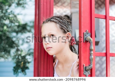 A sad little girl with afro-braids looks out from an old English telephone booth. Generational contrast