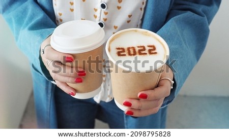 Happy New Year 2022 food art theme female hands holding takeaway disposable double wall paper coffee cups contained cappuccino with number 2022 on frothy surface and another one with plastic lid. Royalty-Free Stock Photo #2098795828