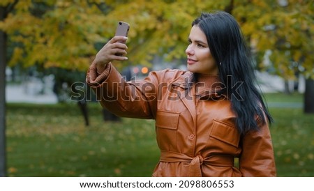 Close-up young hispanic woman stands in autumn park holding phone in hand girl blogger posing photographed on mobile camera make foto for blog using smartphone lady vlogger makes online streaming