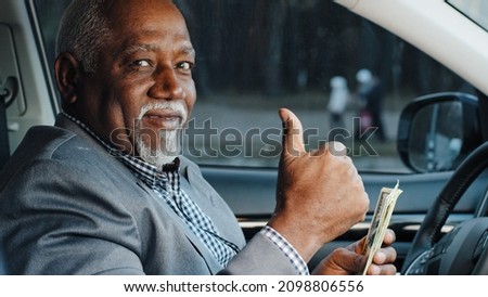 Elderly male sitting in car counting cash payment wages mature african american businessman calculates income successful business happy smiling man looking at camera shows thumbs up gesture approval Royalty-Free Stock Photo #2098806556