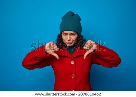 Disappointed upset sad frustrated pretty woman in bright red coat and green hat looking at camera and showing thumbs up, posing against blue background with copy ad space