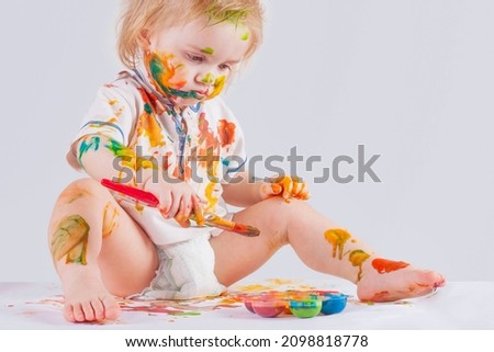 The baby is focused. The child messy paints his face and clothes with paint. 