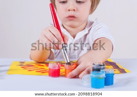 Child draws with paints. Big red paintbrush in a hand. Chooses orange