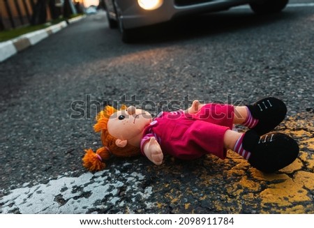 A sad plush toy doll is lying on the road near the wheel of a car. The concept of a road traffic accident. Royalty-Free Stock Photo #2098911784