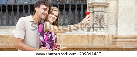 Panoramic portrait of an attractive young couple relaxing taking a selfie picture of themselves with a smartphone while visiting a destination city on holiday, together outdoors. Technology lifestyle.