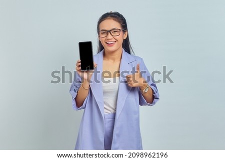 Portrait of cheerful young Asian woman showing blank screen mobile phone and gesturing thumbs up isolated on white background