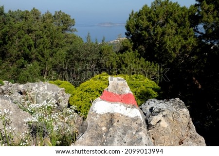 Sign of the Caria Way painted on rocks in Bodrum. Long distance trail sign with two stripes, one red and one white, trekking sign with blurred mountains and sea in the background.