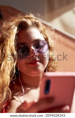 Caucasian girl with blonde hair leaning on a wall looking at the phone with sunglasses