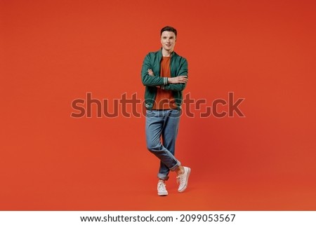 Full size body length smiling vivid happy young brunet man 20s wear red t-shirt green jacket hold hands crossed isolated on plain orange background studio portrait. People emotions lifestyle concept Royalty-Free Stock Photo #2099053567