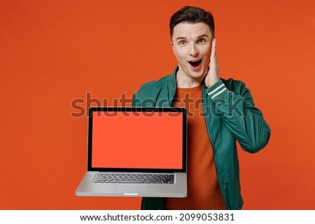 Shocked fun young brunet man 20s wears red t-shirt green jacket hold use laptop pc computer with blank screen workspace area keeping mouth wide open isolated on plain orange background studio portrait