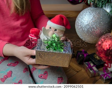 A girl sitting on the floor by the Christmas tree and holding a small Christmas present and a Santa Claus toy in her hands