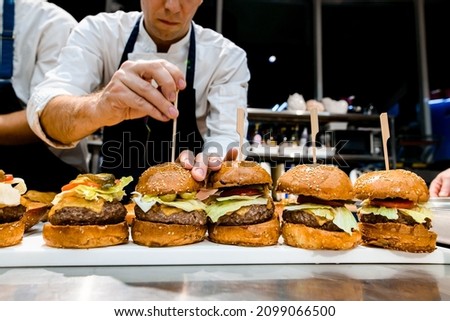 front view of delicious juicy hamburgers with meat cutlet and vegetables on the table. Hand of chef pierces burger with wooden stick