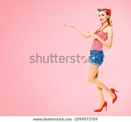Full body of smiling woman pointing at something. Excited girl in pin up, showing, holding advertise some product or copy space. Retro fashion and vintage. Rose pink color back. Adviser advisor advert