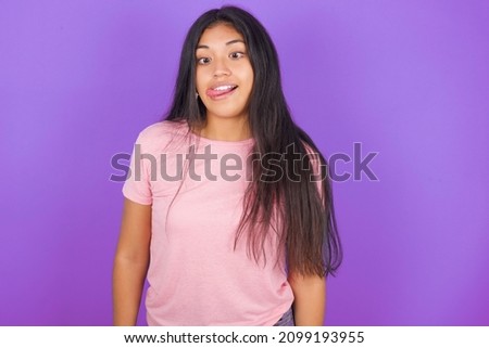 Funny Hispanic brunette girl wearing pink t-shirt over purple background  makes grimace and crosses eyes plays fool has fun alone sticks out tongue.