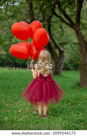 a cheerful  girl stands in a green apple park in a red fluffy dress and holds a lot of balloons in the form of hearts in her hand, a cheerful child indulges in a holiday and shows positive emotion