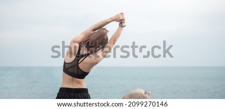 Young fitness female in sportswear stretching body against ocean view, healthy woman exercise in morning. Workout, wellness and work life balance concepts