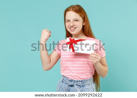 Little redhead kid girl 12-13 years old in pink striped t-shirt hold gift voucher flyer mock up do winner gesture clench fist isolated on pastel blue background. Children lifestyle childhood concept