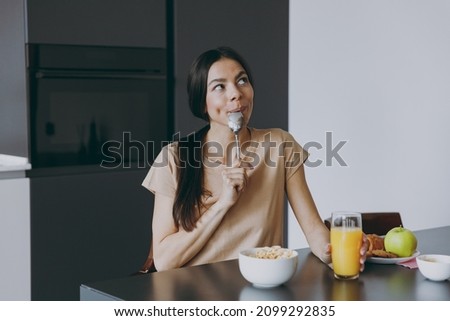 Young happy dreamful minded housewife woman 20s wearing casual clothes beige shirt eat oatmeal porridge muesli in morning cooking food in light kitchen at home alone. Healthy diet lifestyle concept. Royalty-Free Stock Photo #2099292835