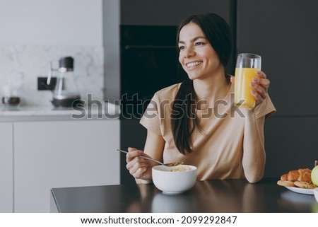 Young minded brunette housewife happy woman 20s wearing casual clothes beige shirt eat oatmeal porridge muesli drink juice cooking food in light kitchen at home alone. Healthy diet lifestyle concept Royalty-Free Stock Photo #2099292847