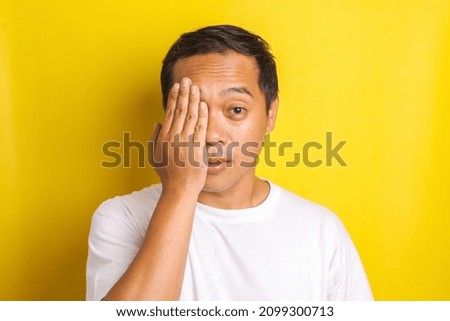 Close-up portrait of Asian man covering his half face with hand with one eye looking to camera isolated on yellow background
