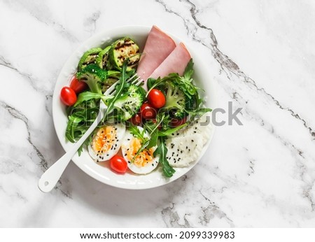 Delicious balanced breakfast, snack bowl - ham, boiled egg, grilled zucchini, green salad in one bowl on a light background, top view       