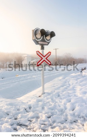 Level crossing sign at a crossing with snow-covered railroad tracks and road and barely visible. Sunny morning after snowfall. Background of trees and buildings. Winter concept