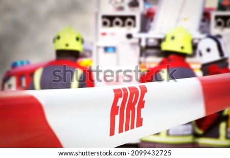 Scene of incident is cordoned off with red and white tape, reading ‘fire’. Out of focus in the background are three firefighters, two fire engine appliances and smoke billowing through the air. Royalty-Free Stock Photo #2099432725
